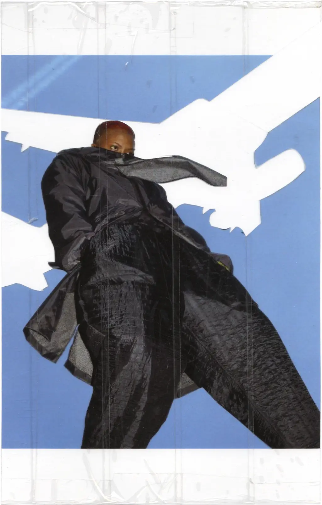 Image of a person taken from very low to the ground, framing them against the blue sky. A low flying airplane encompasses most of the background. The entire image and the acetate on which it was printed is neatly covered with rows of clear packaging tape. The outline of the airplane has been crudely cut out of the transparent tape with a box cutter which allowed the alcohol wash to completely remove it while leaving the person and the sky untouched.